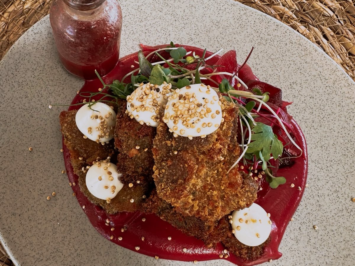 A plated dish of crumbed pieces on a bright beetroot purée, topped with cream, puffed grains and microgreens, with a small bottle of sauce beside it